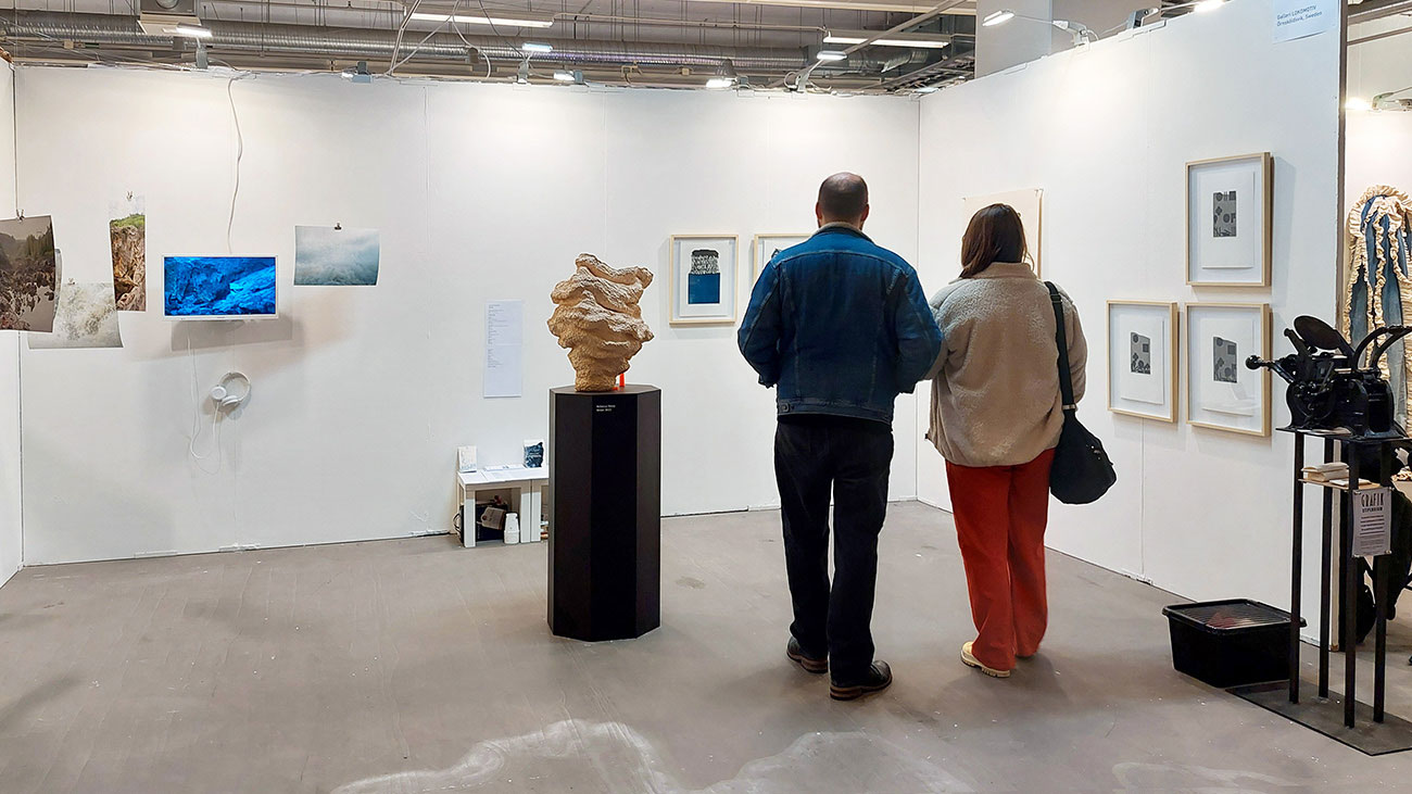 Foto. En monter med vita väggar där det hänger bilder. Mitt på golvet står ett podium med en skulptur. En man och en kvinna står med ryggen mot kameran och tittar på konsten. 