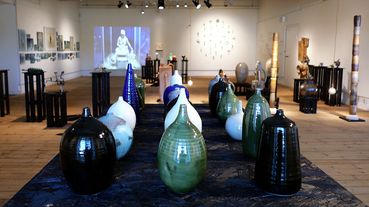 En vy över utställningen. I förgrunden står flera keramikvaser i bl.a. grönt, vitt blått och svart. I bakgrunden syns skulpturer, videoprojektion och andra verk. 