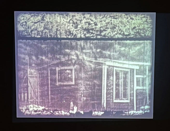 Stillbild ur film. På bilden syns ett hus, lite diffust. Bilden ser gammal, repig och nött ut i blek gråskala. 