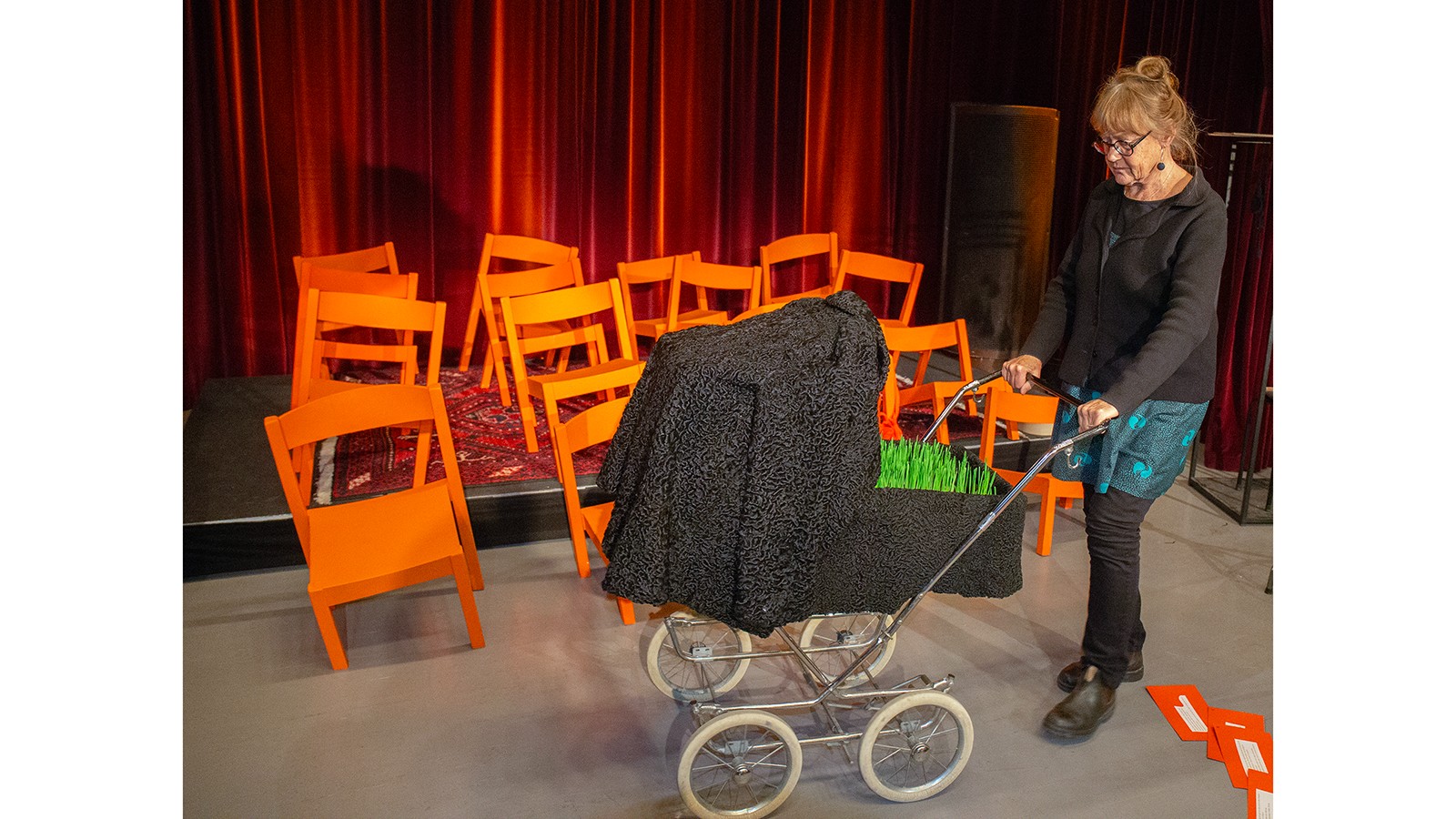 En kvinna med uppsatt hår, glasögon och mörka kläder, står med en barnvagn som det växer gräs i. Hon står framför 16 orangea stolar med ojämna ben. 