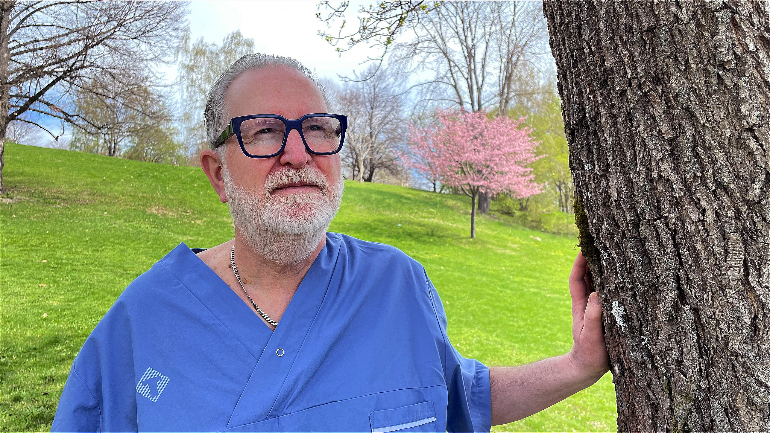 Manlig tandläkare står utomhus och har en hand på ett träd.