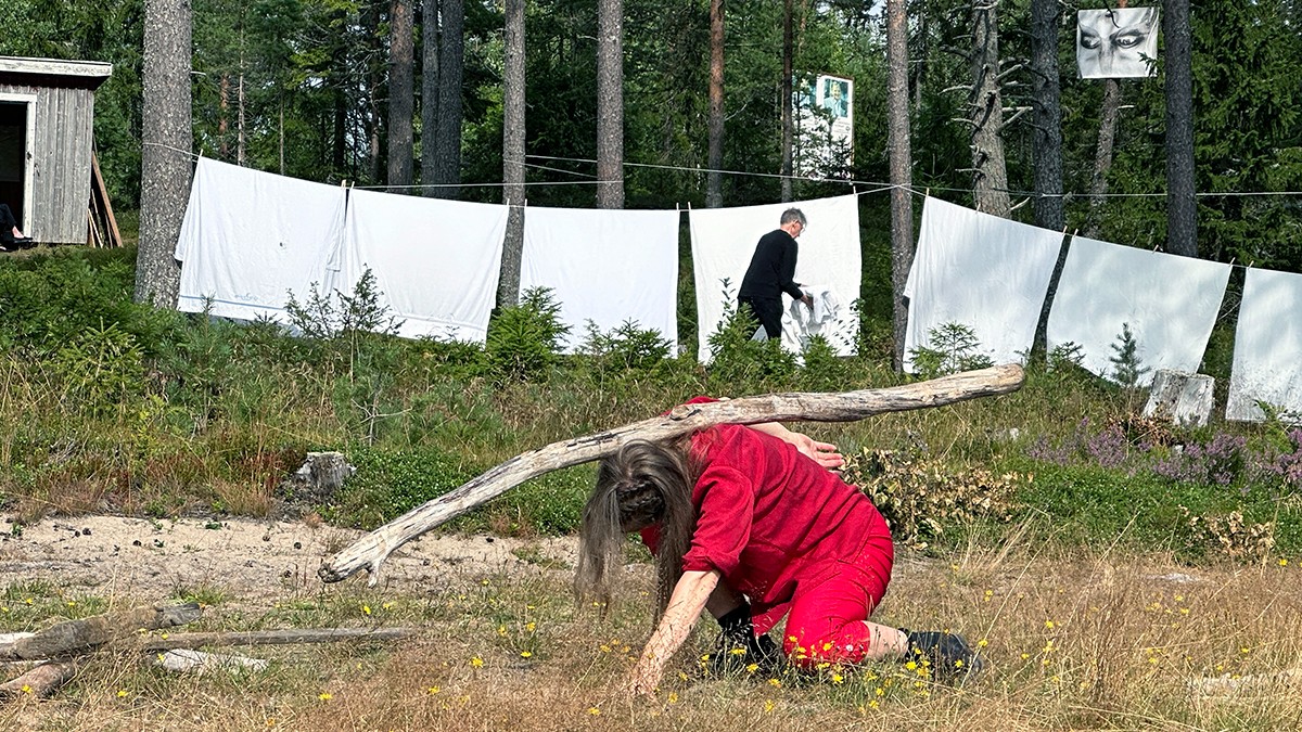 En kvinna klädd i rött dansar med en lång bit drivved på en öppen yta. I bakgrunden syns en man klädd i svart hänga tvätt, vita lakan, mellan träden. 