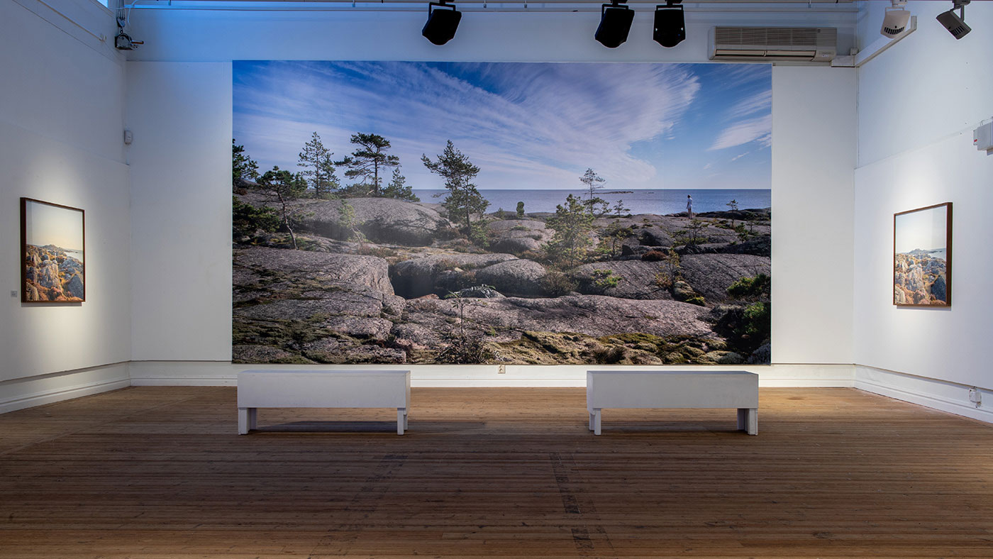 Vy från utställningen. Ett rum med trägolv och vita väggar. Mitt i rummet står två vita bänkar bredvid varandra. Framför bänkarna på väggen rakt fram hänger ett stort fotografi. Det täcker nästan hela väggen. Fotot föreställer ett kostlandskap, med klippor och växtlighet bestående av små barrträd och ljung. Längre bort syns havet och horisonten ligger en bit ovan bildens mitt. Himlen är blå med molnformationer som sträcker sig bortåt. 