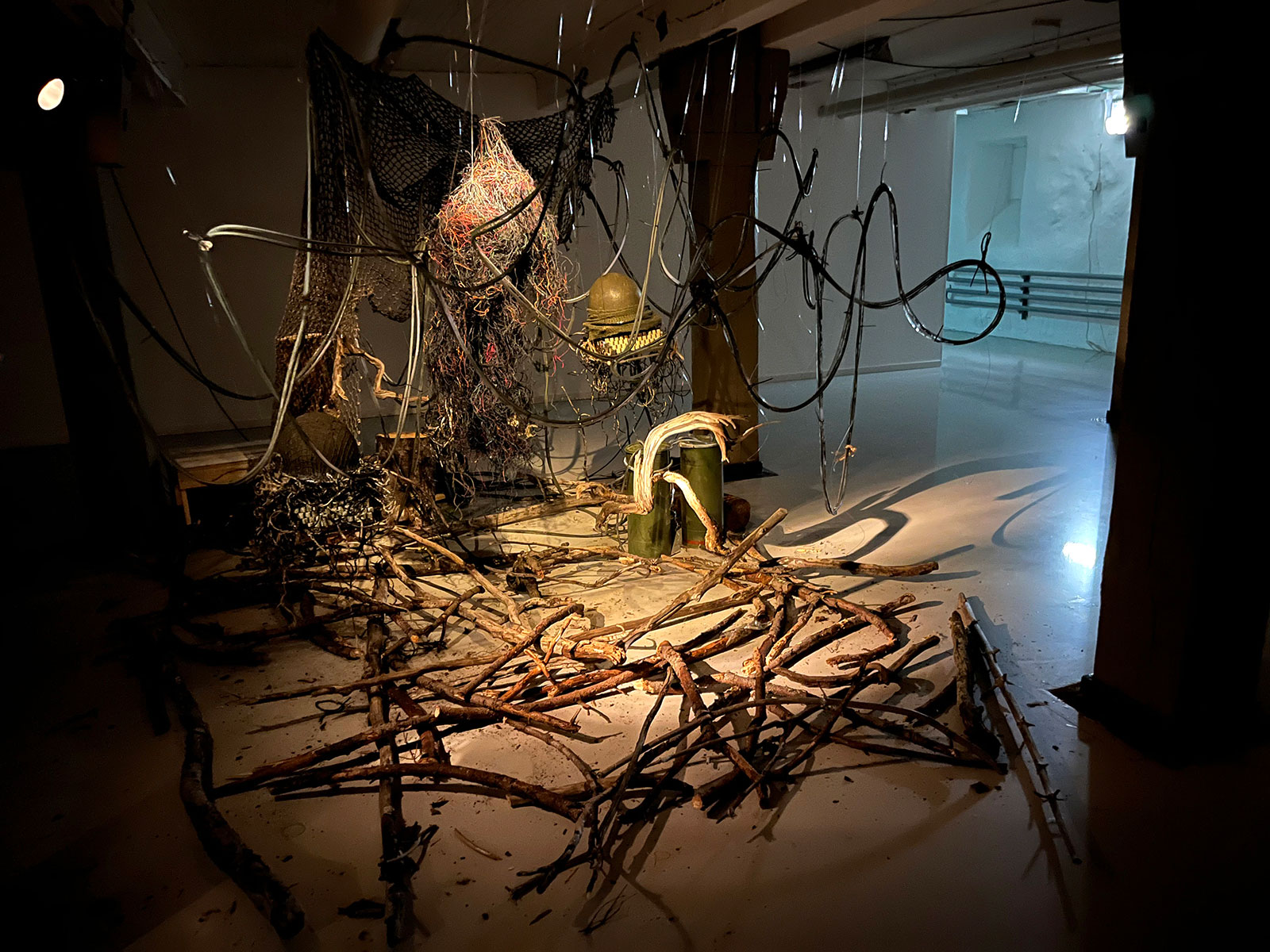Foto av installation. I mitten av ett rum, delvis dunkelt upplyst, ses en stor trasslig hopsättning av föremål såsom trädgrenar, gamla elkablar, skrivmaskiner och militärhjälmar. 