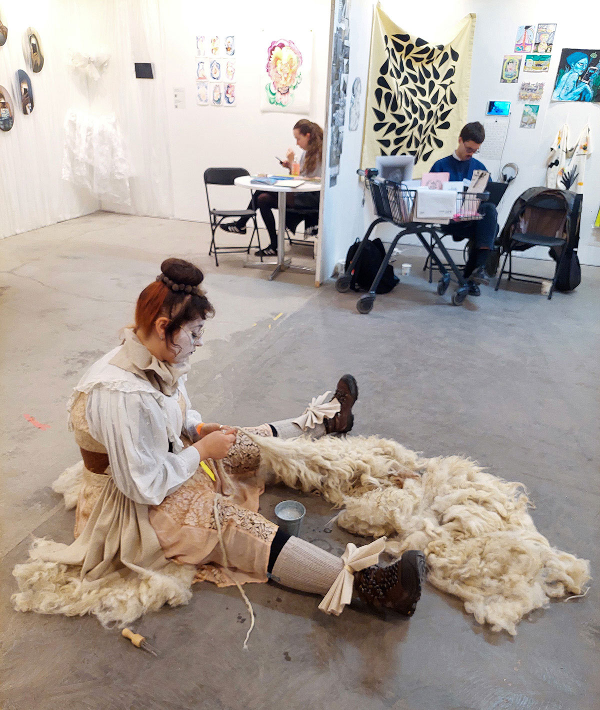 Foto. En kvinna med vitsminkat ansikte och rödaktigt uppsatt hår sitter på golvet och arbetar med vad som ser ut som en fårskinnsfäll. I bakgrunden syns personer sittandes i montrar med konst på väggarna. 