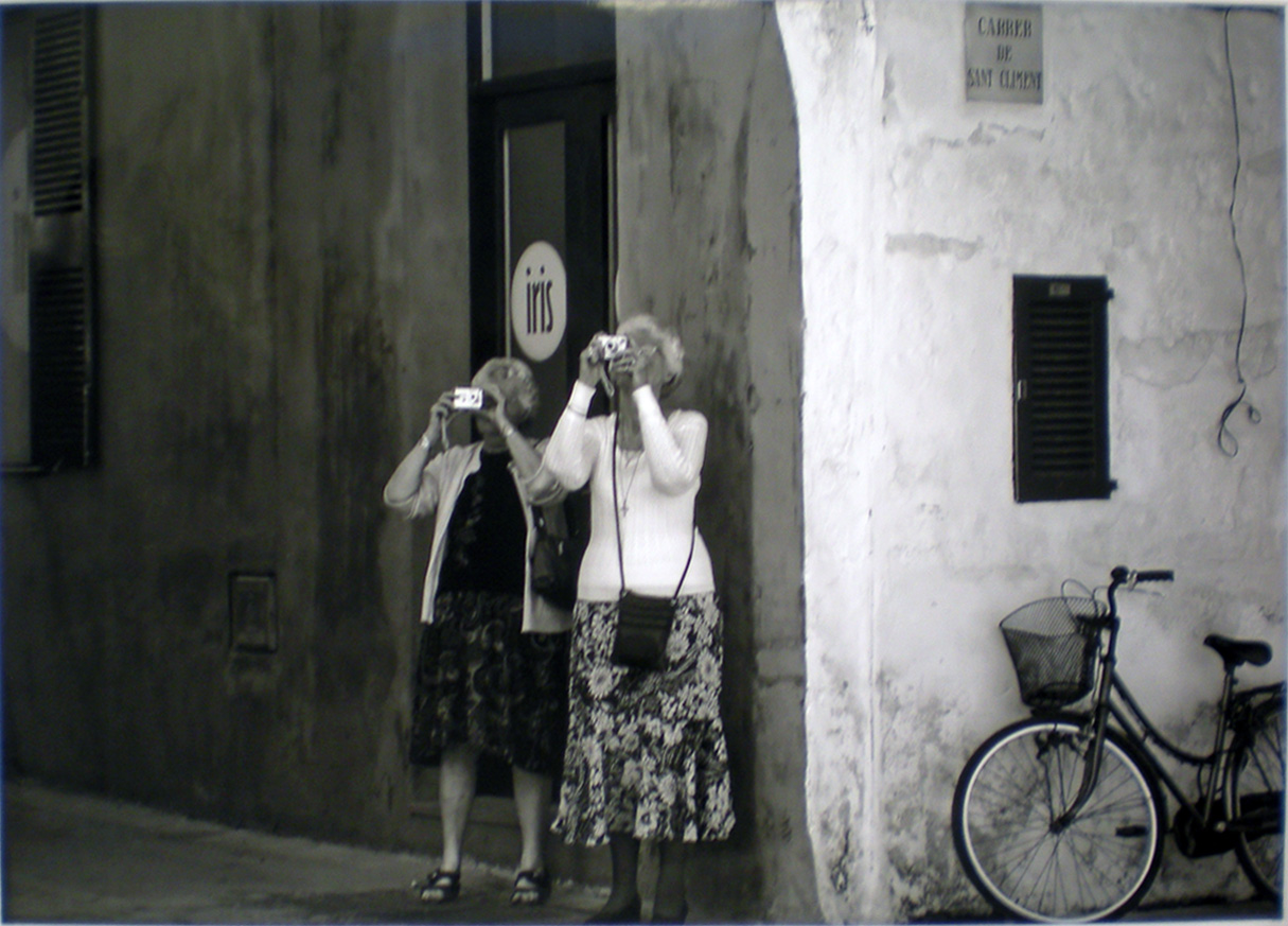 Ett svartvitt fotografi med två äldre damer som står vid ett hörn av ett äldre betonghus. Runt hörnet, till höger, står en cykel med cykelkorg. Ovanför den en trälucka och högre upp en sliten kvadratisk gatuskylt med texten ”Carrier de Sant Clement”. Kvinnorna har båda ljusa koftor. Den ena bär en blommig kjol, till detta en liten axelremsväska, den andra kvinnan bär mörk klänning. Bakom dem en hög trädörr med en rund skylt på med texten ”iris”. Båda damerna står med små kameror framför ögonen och fotar någonting på högre höjd. Gaveln med cykeln är ljus, den andra mörk.