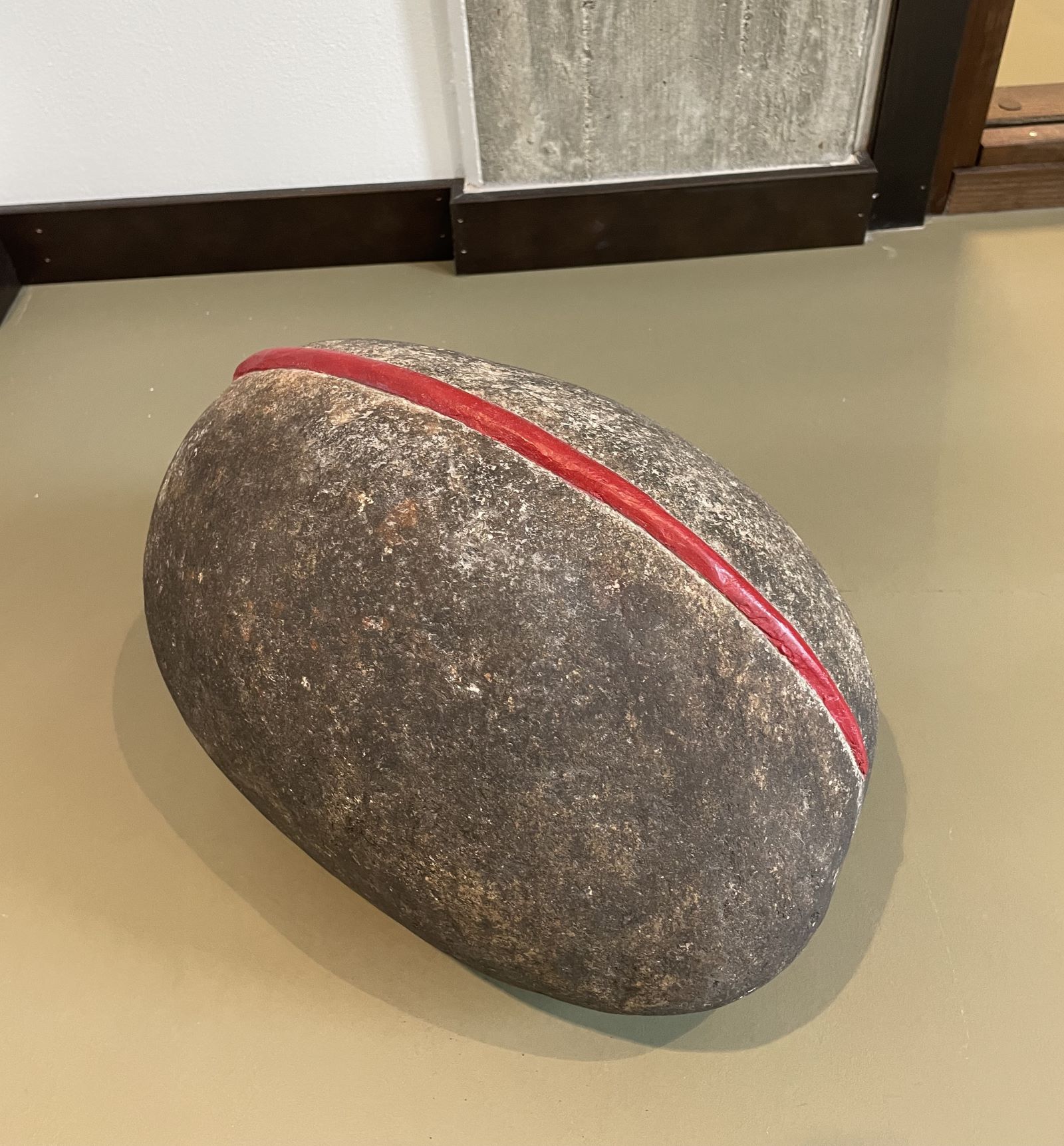 Foto: En oval sten, mjukt rundad. Men på översidan, på längden, en skåra. En huggen fördjupning som är färgad röd. 