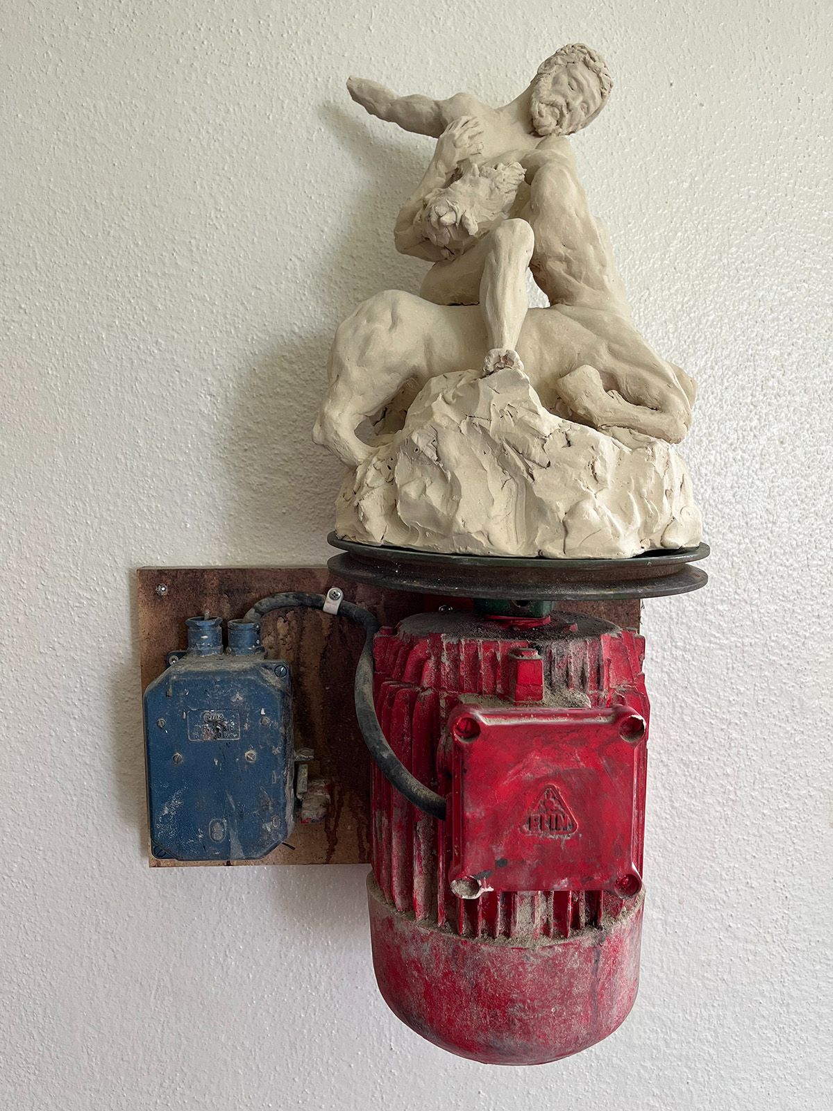 Foto. På en vit vägg hänger en skulptur. En röd cylindrisk del av en motor, från den går en kabel till en blå dosa direkt till vänster. Ovanpå den röda delen sitter en mer klassisk skulptur av en man och en kentaur som verkar strida. 