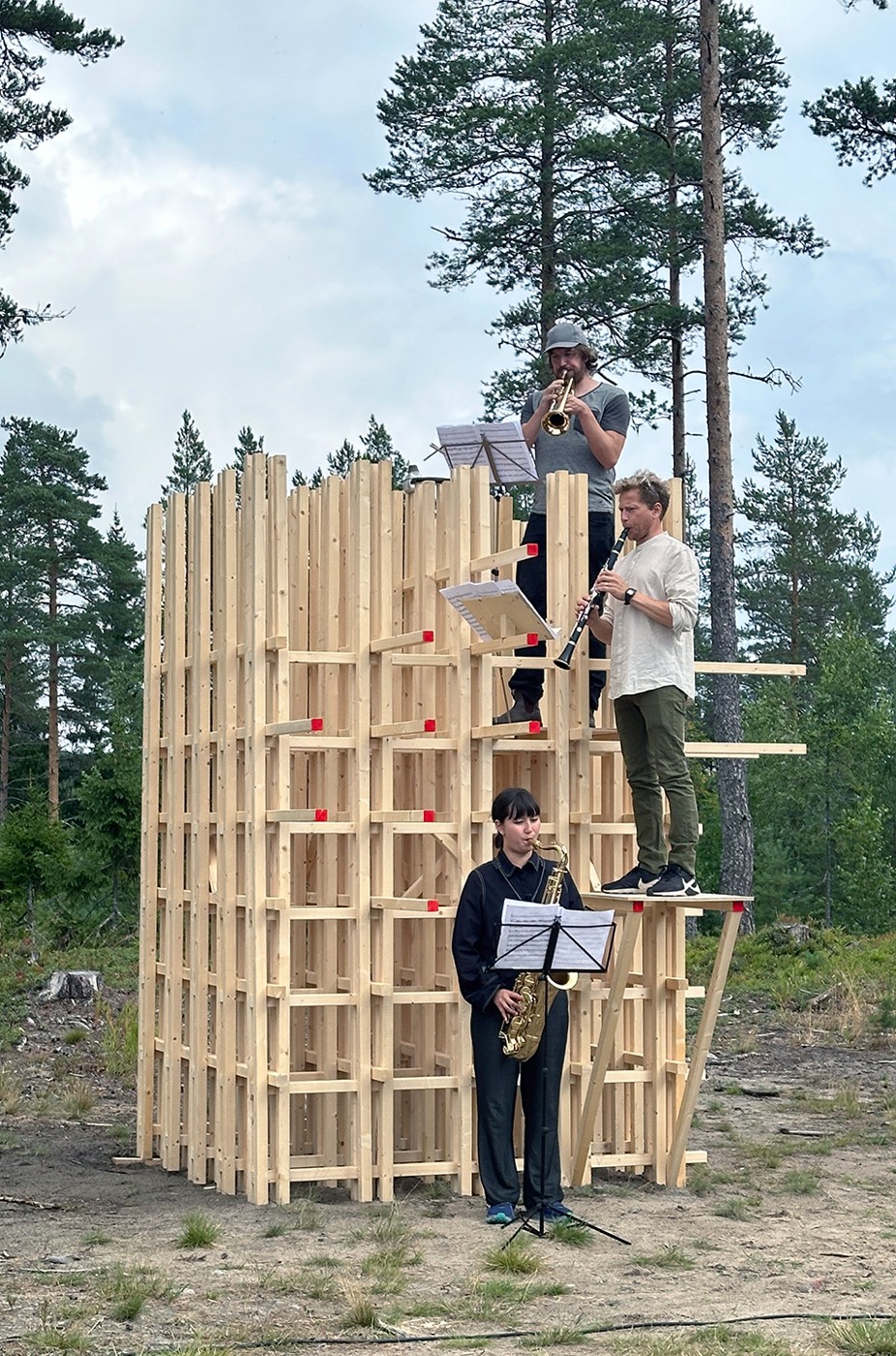 Framför en träkonstruktion står en person och spelar saxofon. Halvvägs upp i konstruktionen, på en avsats, står en andra person med en klarinett. Lite längre upp står en tredje person och spelar trumpet. I bakgrunden syns skog. 