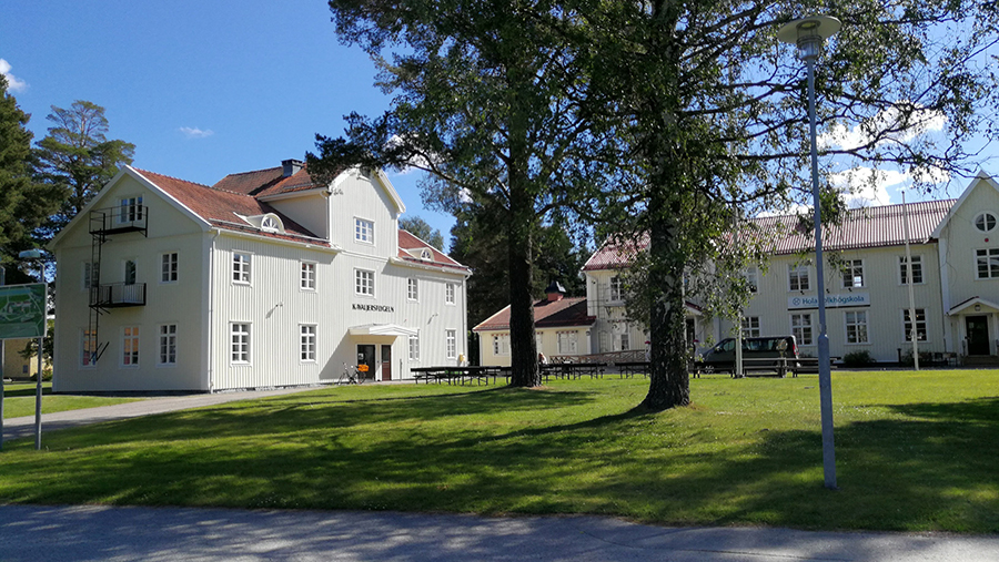 Två grå trähus med en blå himmel som bakgrund. De är omgivna av en grön gräsmatta. Några träd står i förgrunden. 