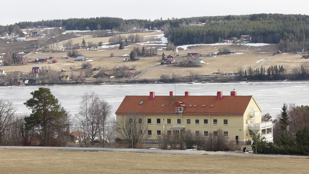 alt='Foto av en lång vit byggnad med tegeltak framför vatten. '