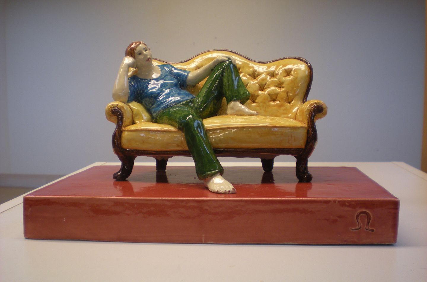 Foto av en skulptur. En gul rokokosoffa på en röd sockel. I soffan sitter en kvinna tillbakalutad med gröna byxor och blå tröja. 