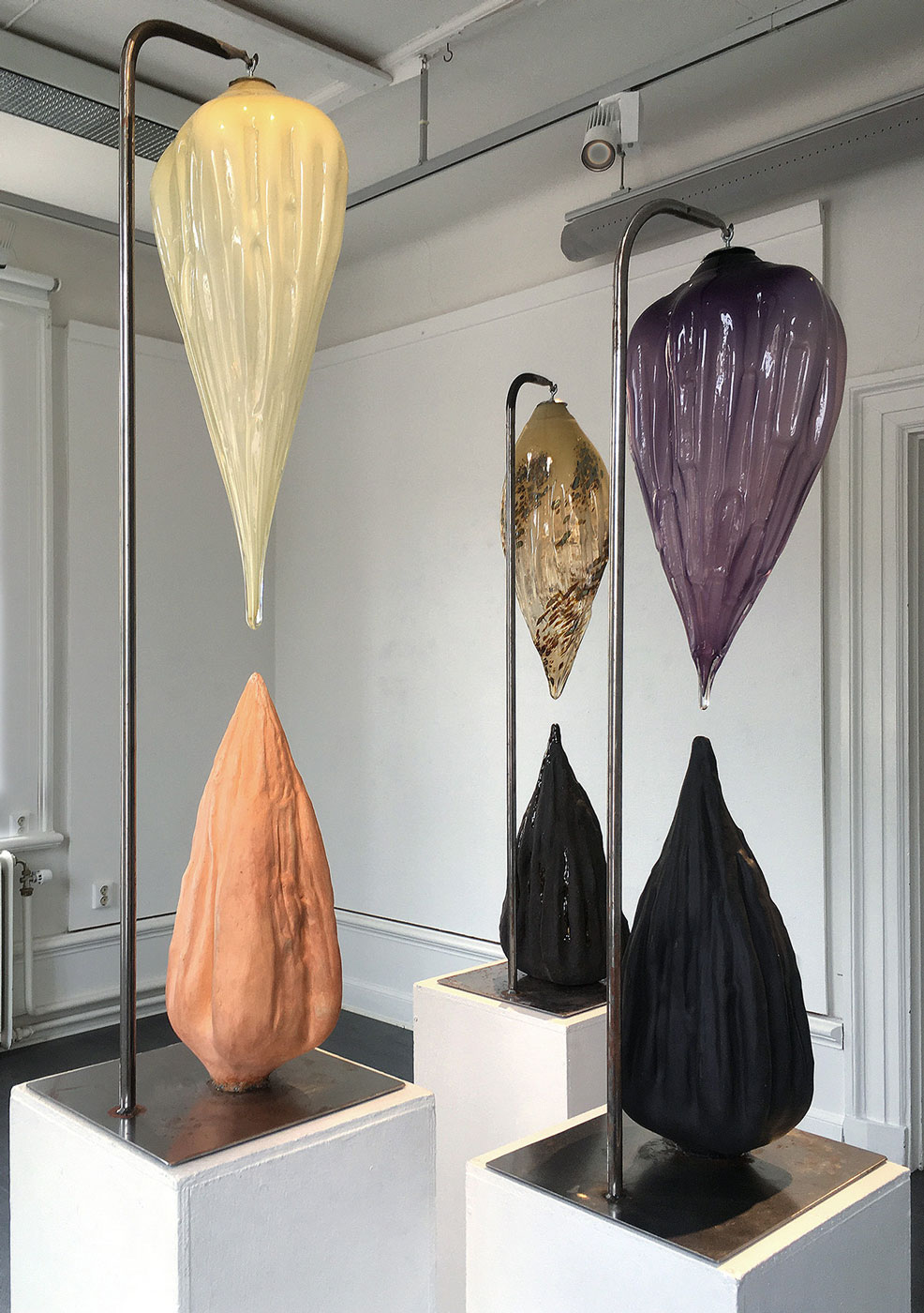 Foto: Tre liknande verk i olika färger; Från en metallkonstruktion hänger en glasform liknande en upp och ner-vänd droppe. Under den en motsvarande form i keramik. 