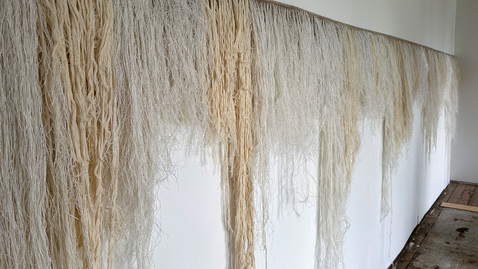 Längs en jämn linje flödar garntrådar, en handvävd rya, i ljusa beigevita färger i olika längder ner längs en vit vägg. 