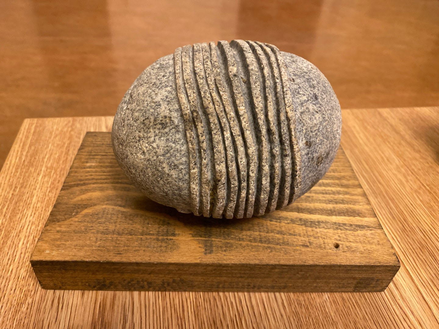 Skulptur i forma av en grå sten på en platta av trä. Stenen har tio uthuggna ränder som går runt dess form. Placerade tätt på mitten av stenen. 