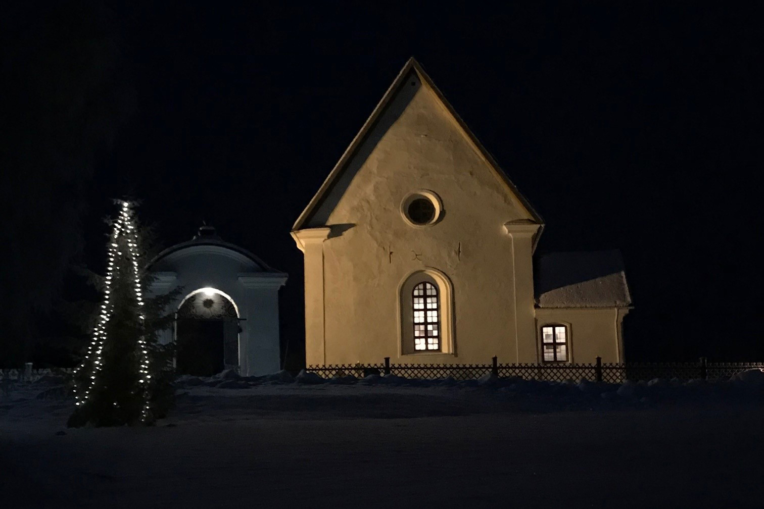 En vit kyrka i ett vinterlandskap med mörk himmel och en gran med ljusslinga.