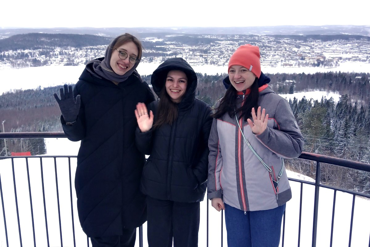 Tre kvinnor står på en balkong med ett vinterlandskap bakom sig och tittar glatt in i kameran.