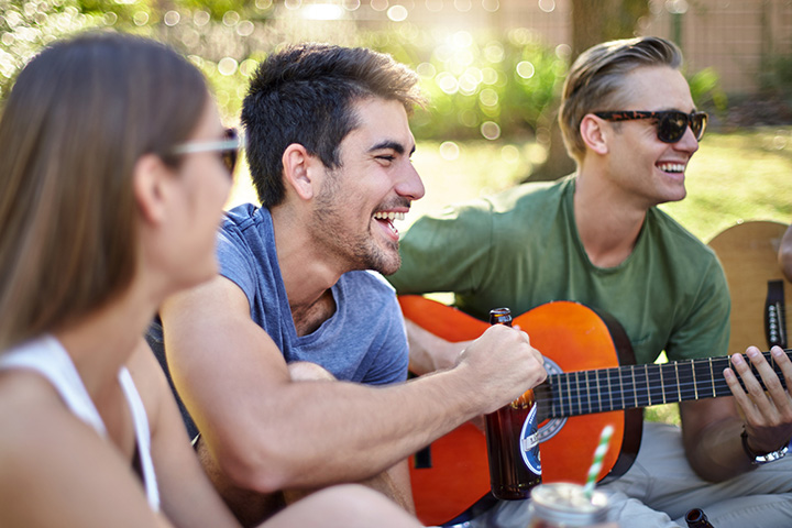 Tre deltagare sitter utomhus och sjunger och skrattar medan en av dem spelar gitarr.