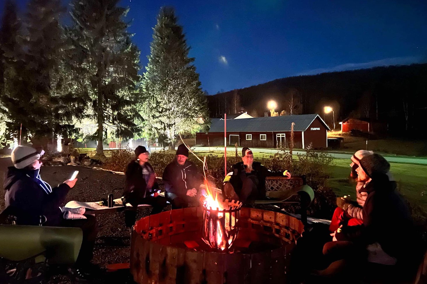 En grupp personer sitter runt en stor lägereld under en mörkblå himmel.