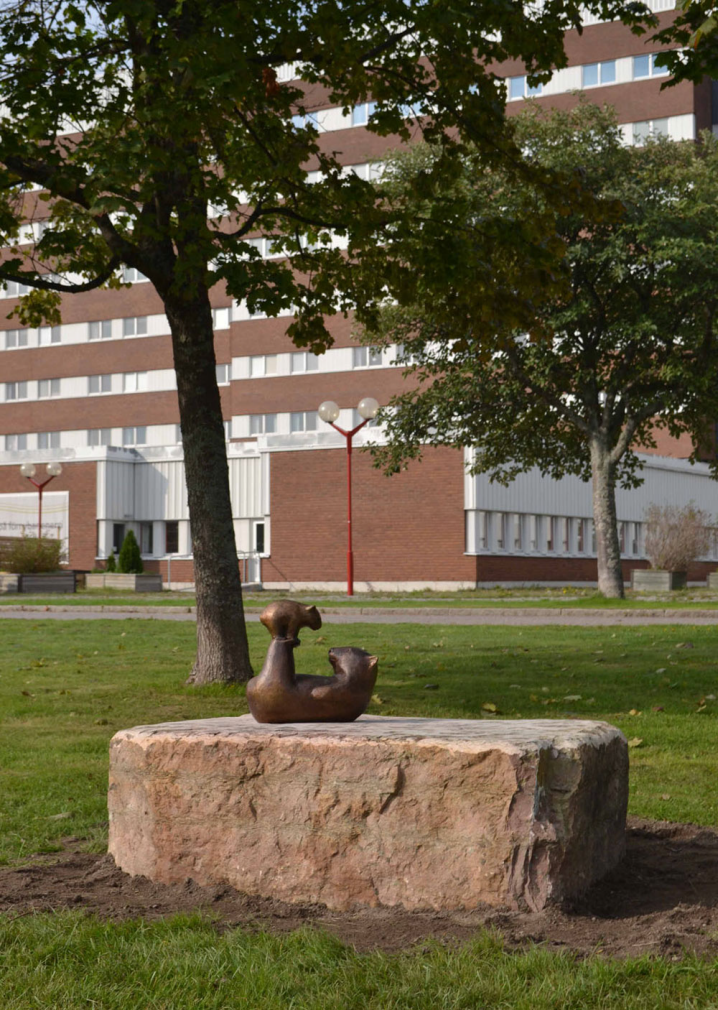 Skulptur, placerad utomhus under ett par träd, i form av en vombat liggande på rygg som har sin unge upplyft på fötterna.